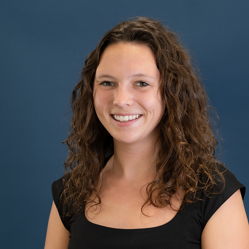 Smiling person with curly brown hair wearing a black top, standing against a plain blue background.