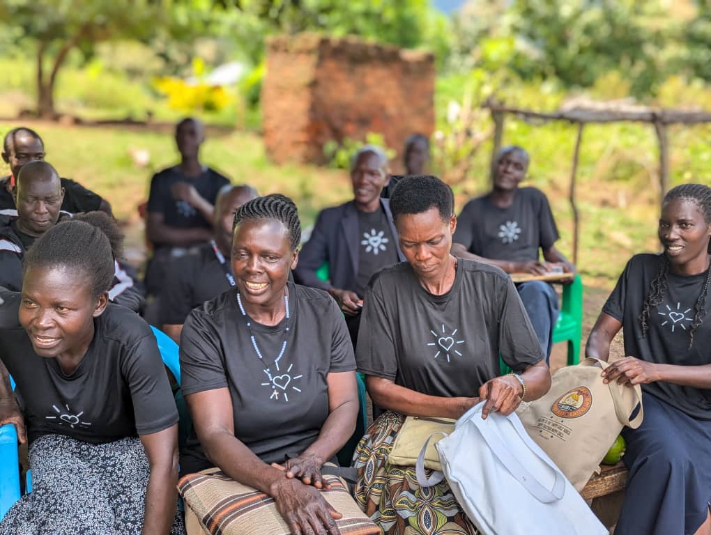 A group of people wearing black shirts with heart designs sit outdoors, some smiling and chatting, with trees and a brick structure in the background.