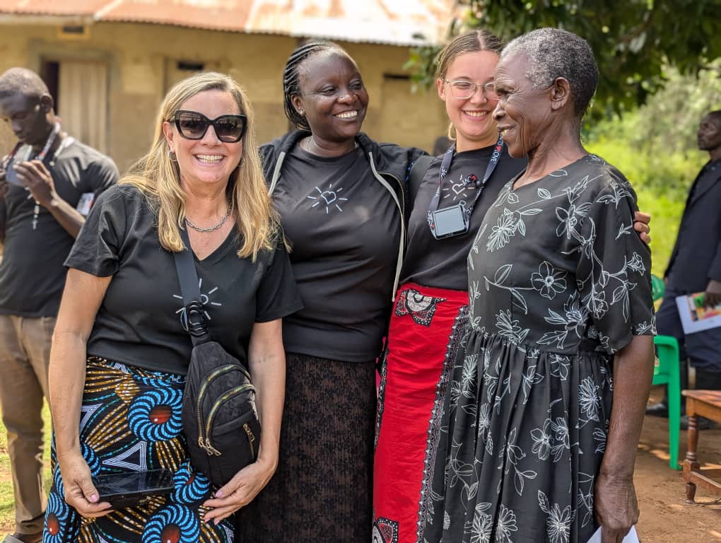Four women in patterned skirts and black tops stand smiling outdoors, with two men in the background. They appear happy and relaxed.