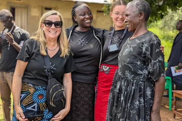 Four women in patterned skirts and black tops stand smiling outdoors, with two men in the background. They appear happy and relaxed.