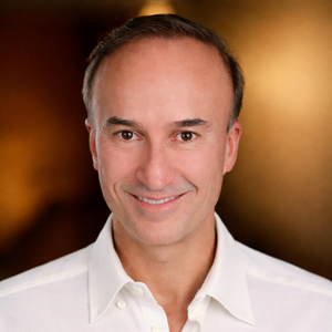 Smiling person wearing a white shirt, standing against a blurred brown background.