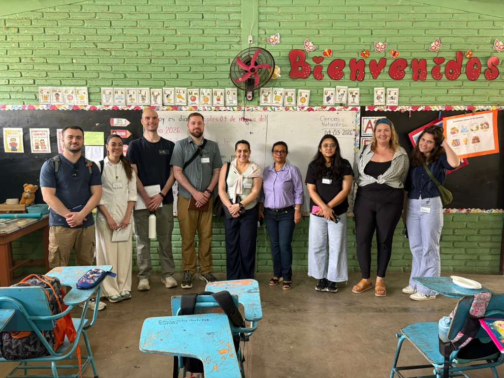 A group of people standing in a classroom in Honduras with a "Bienvenidos" sign above a chalkboard. Desks and chairs are visible in the foreground.