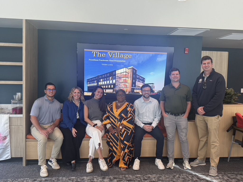 A group of seven people smiling in front of a screen displaying "The Village" with an image of a building. They are seated and standing indoors.