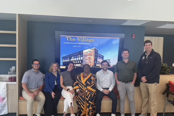 A group of seven people smiling in front of a screen displaying "The Village" with an image of a building. They are seated and standing indoors.