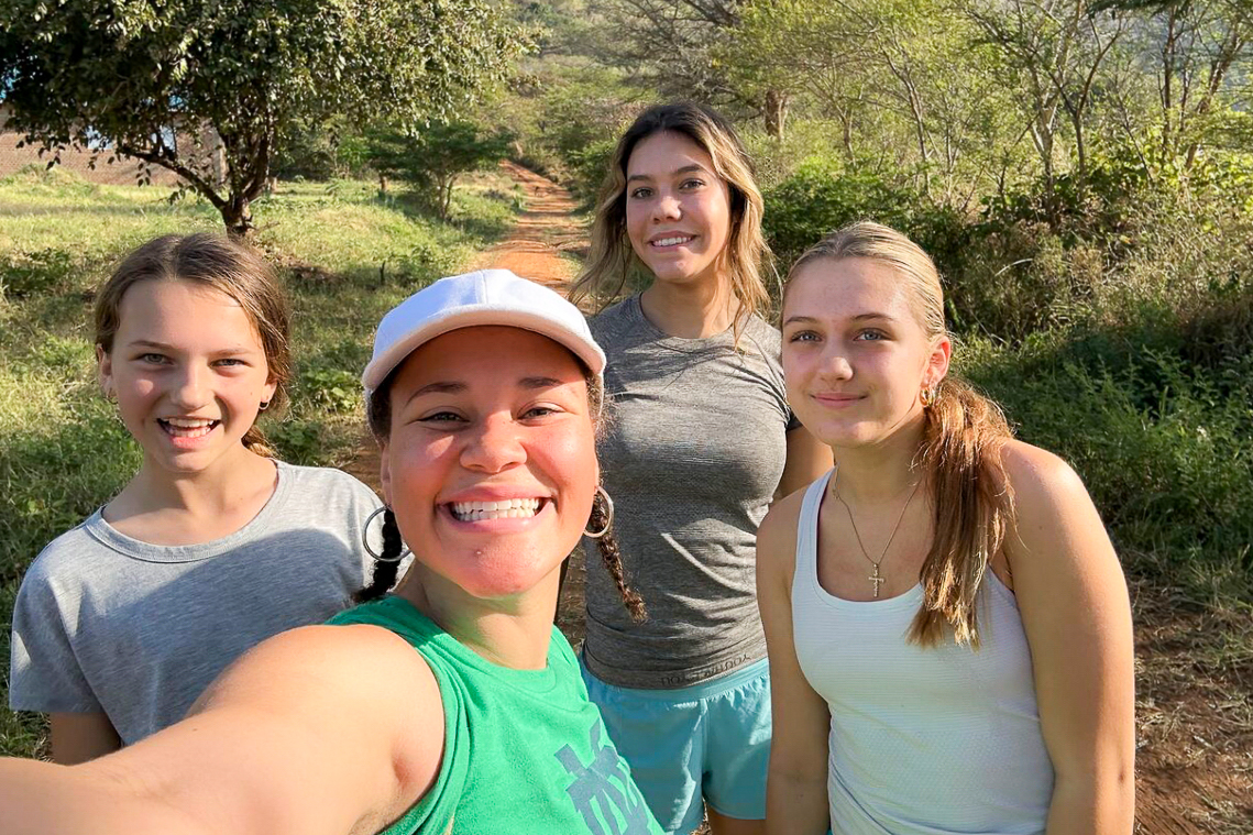 Four people smiling for a selfie on a sunny day, standing on a dirt path surrounded by greenery and trees.