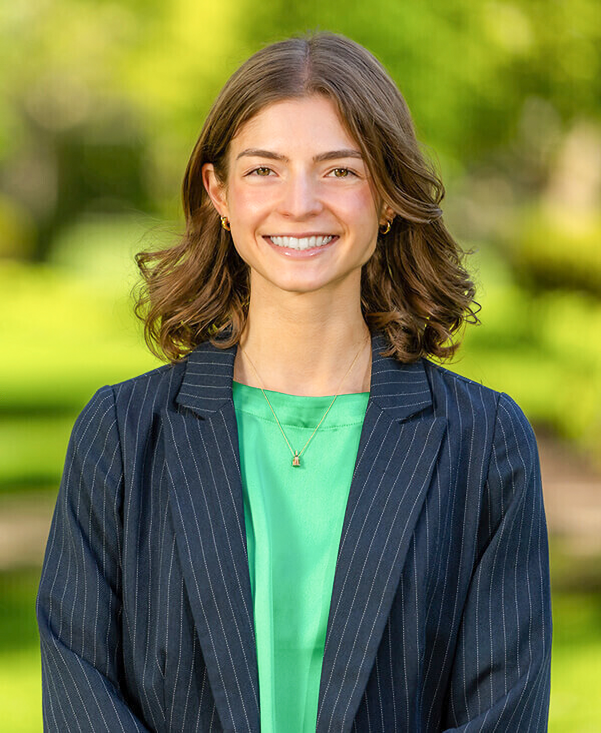 Smiling person with brown hair, wearing a striped blazer and green top, standing outdoors with a blurred green background.