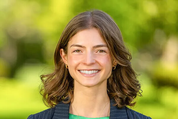 Smiling person with brown hair, wearing a striped blazer and green top, standing outdoors with a blurred green background.