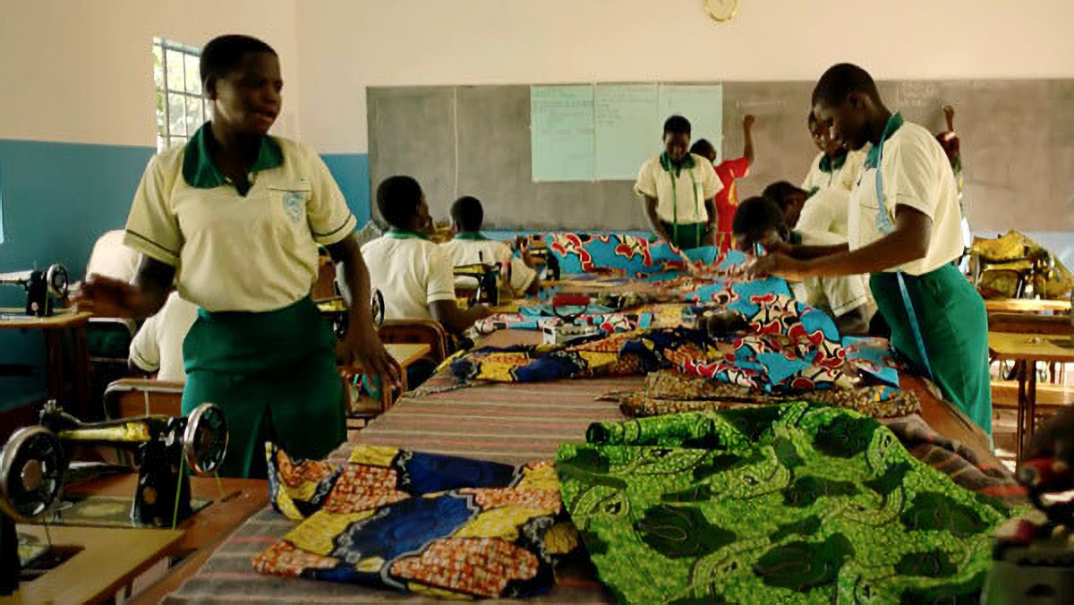 Students in a classroom work with vibrant African fabrics on sewing machines, wearing school uniforms.