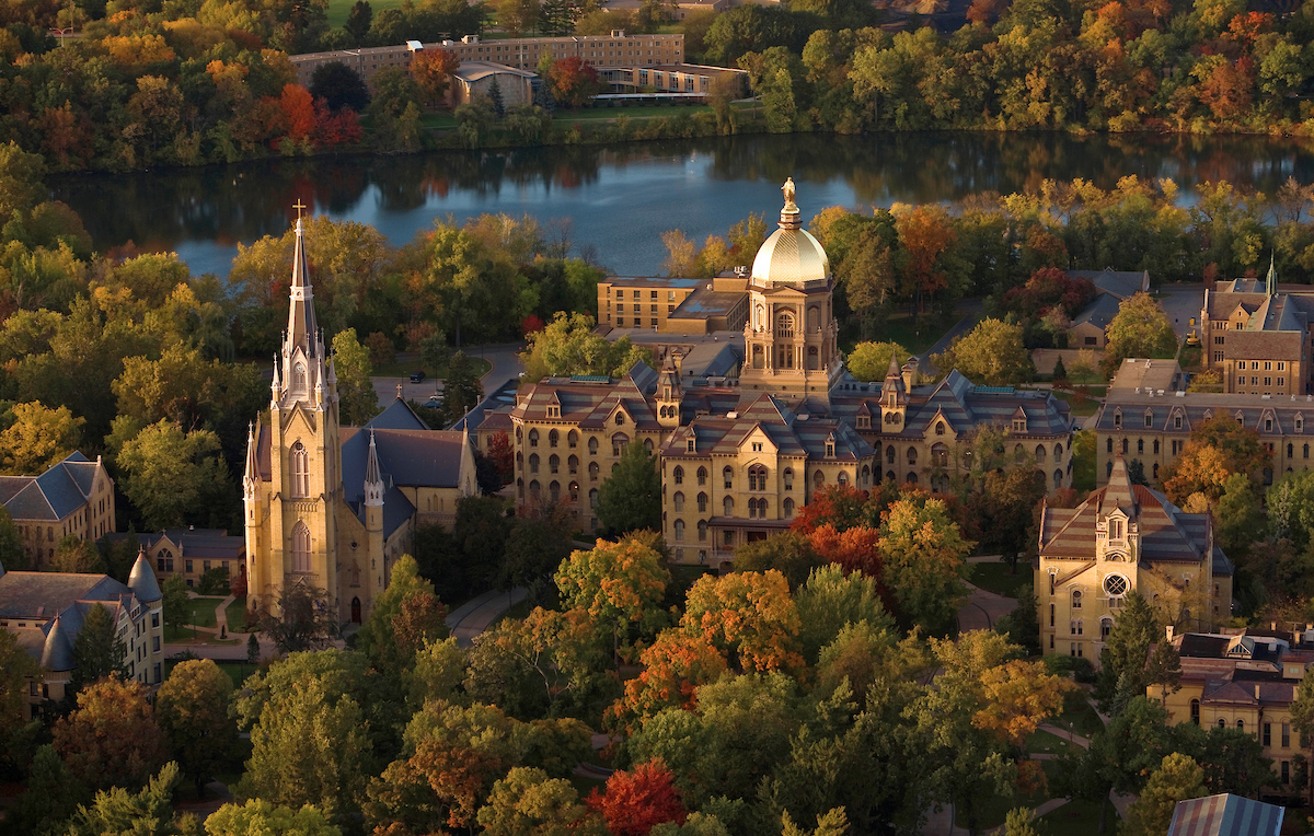 Aerial view of the university of Notre Dame campus with a golden dome and basilica, surrounded by trees and a lake in autumn colors.