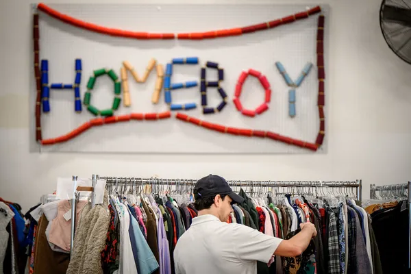 A Frontlines alumni advisor browses through a rack of clothes in Homeboy Threads warehouse.. Above, a colorful sign spells "HOMEBOY" on the wall.