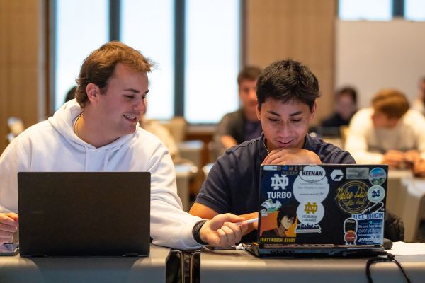 Two people sitting side by side working on laptops covered in stickers, engaging in a discussion in a well-lit room.