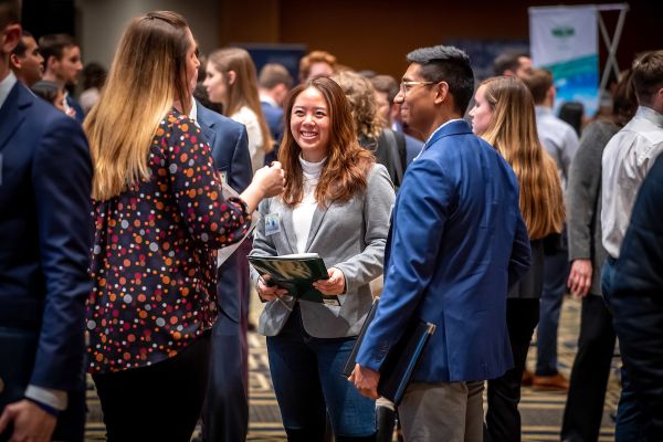 A female student in a grey blazer, a male student in a blue blazer and recuriter in a patterned shirt are standing in a circle in a conversation. They are in a room full of ther students and employers for a career fair with tables and banners in the background.