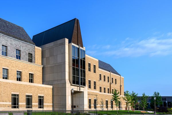 A modern academic building with tan brick walls, large windows, and a sloped roof, set against a clear blue sky.
