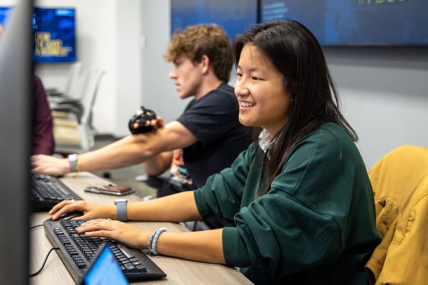 A female student in a green sweater smiles while working on a computer, beside a man using a camera in a classroom setting.