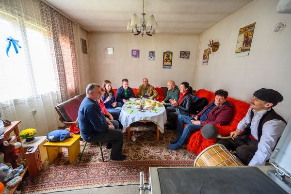 A group of people sit around a table with food in a cozy living room in Zvechan North Macedonia, decorated with posters and a chandelier, engaged in conversation and listening to a man play a percussion instrument.