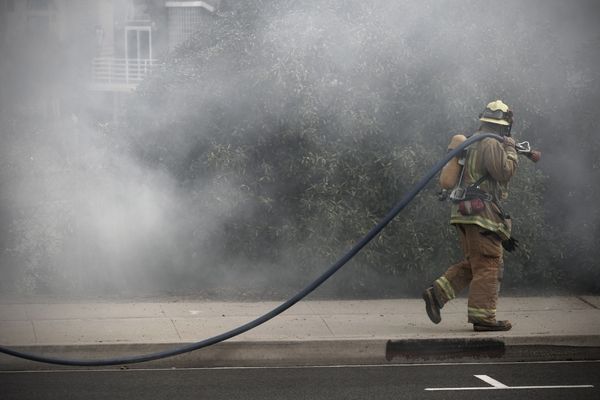 Firefighter in gear walks on a sidewalk, holding a hose, surrounded by smoke, with bushes and buildings in the background.