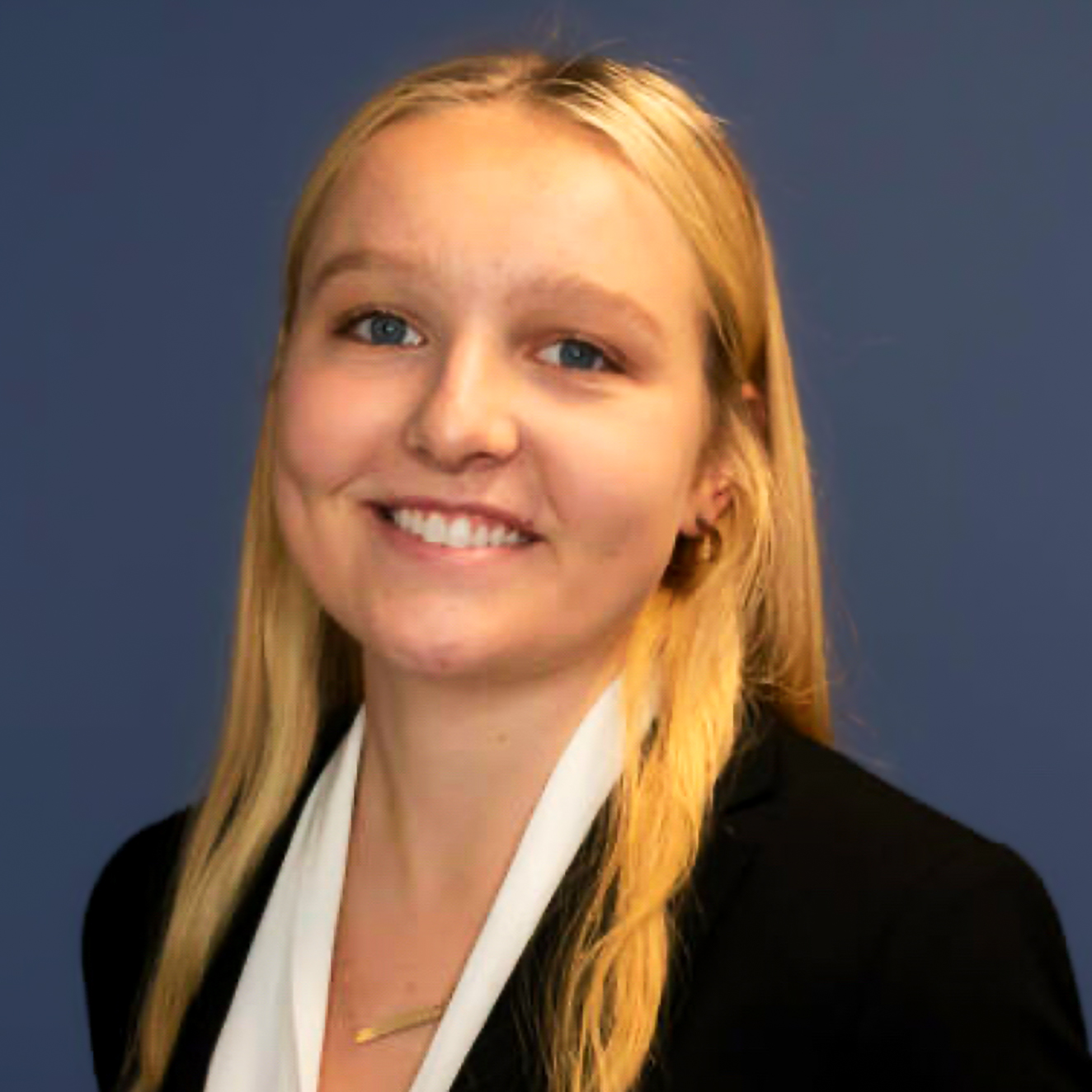 Smiling person with long blonde hair in a black blazer and white shirt against a blue background.