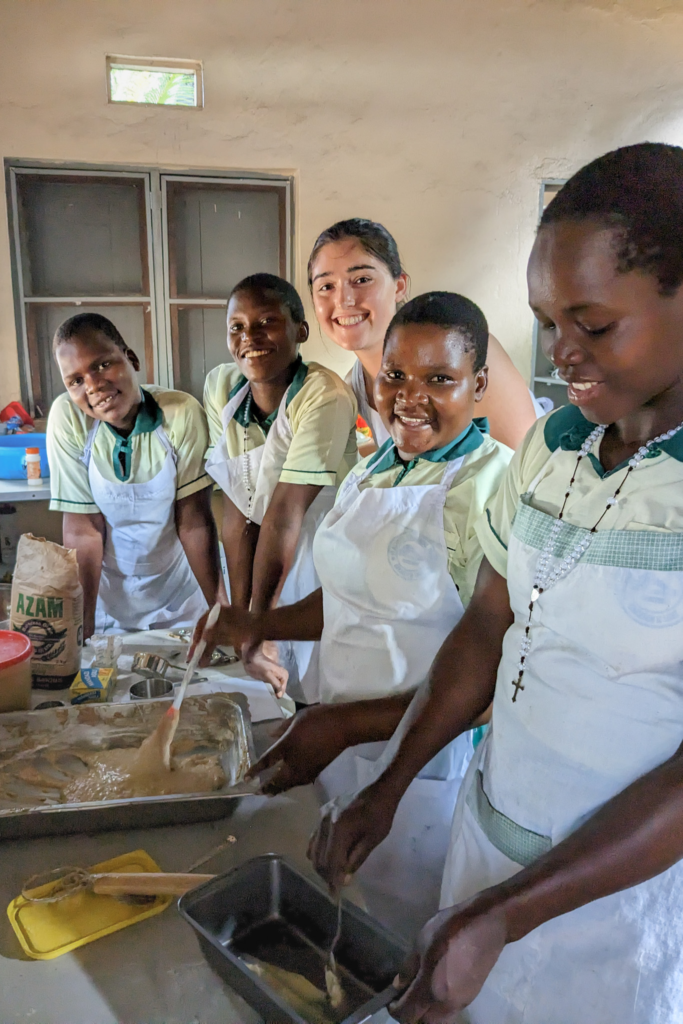 A group of five people, wearing white aprons, smile as they work together to prepare a dish in a kitchen setting.