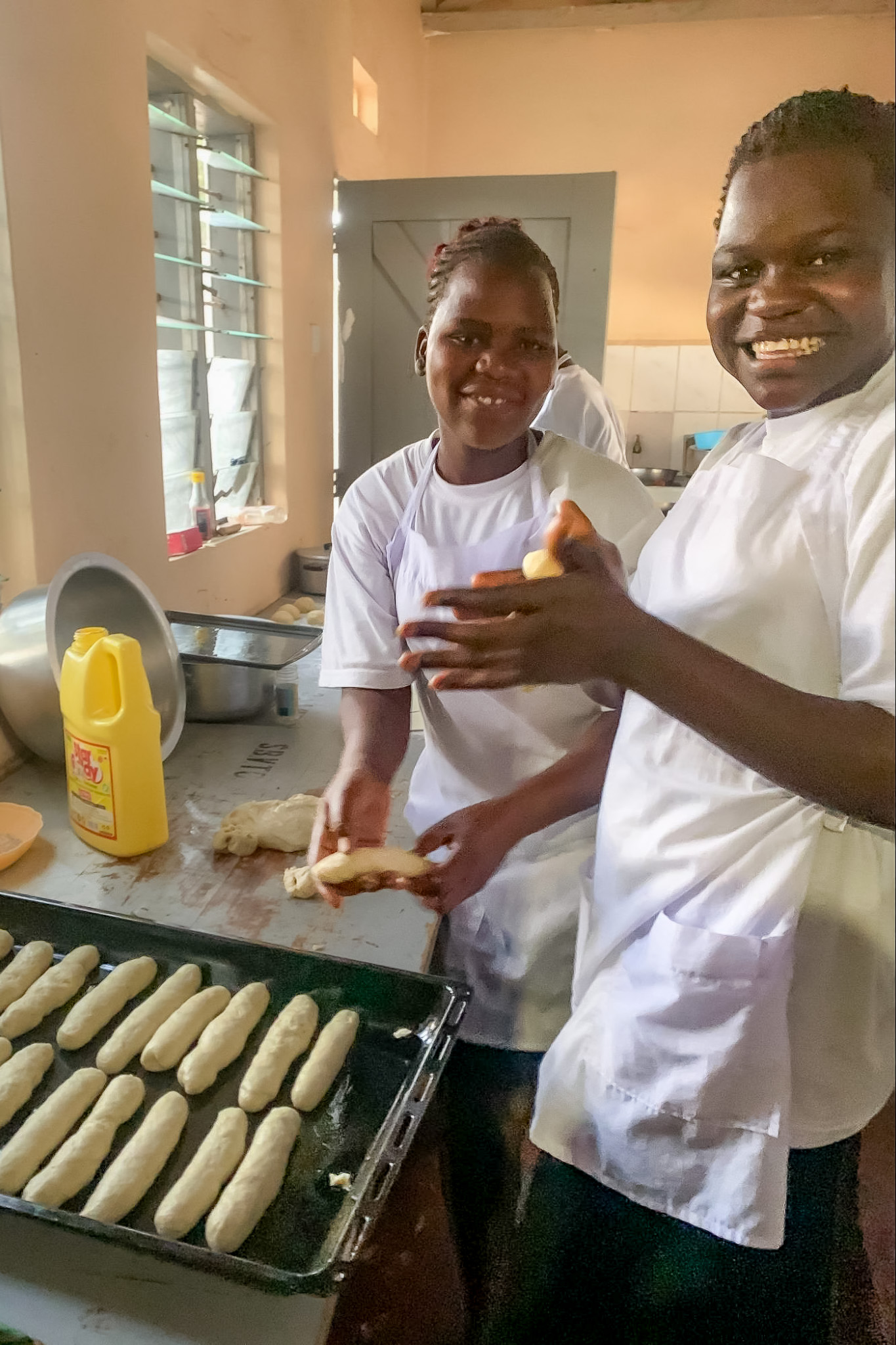 Two smiling people in white aprons prepare dough in a kitchen, placing shaped pieces on a baking tray.