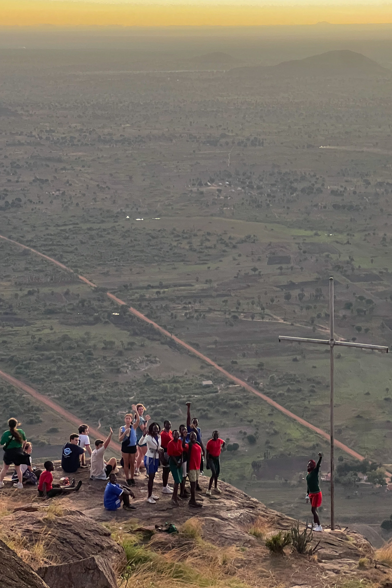 A group of people gathers near a large cross atop a hill, overlooking a vast landscape with a sunset sky.