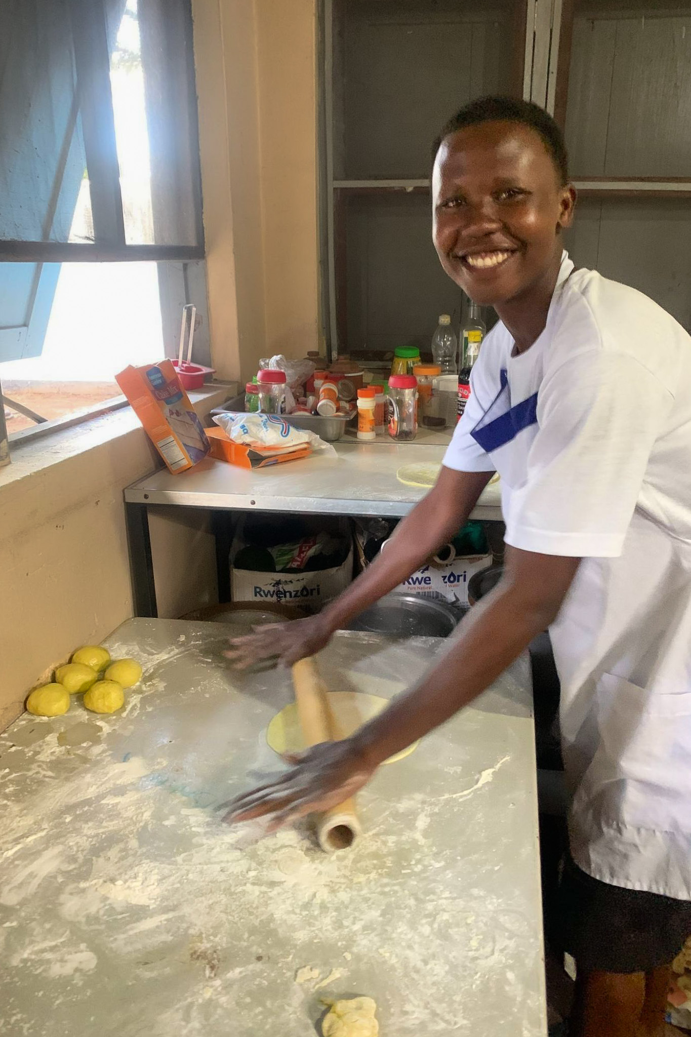 Smiling person rolling dough on a floured table in a kitchen, with ingredients and spices on the counter nearby.
