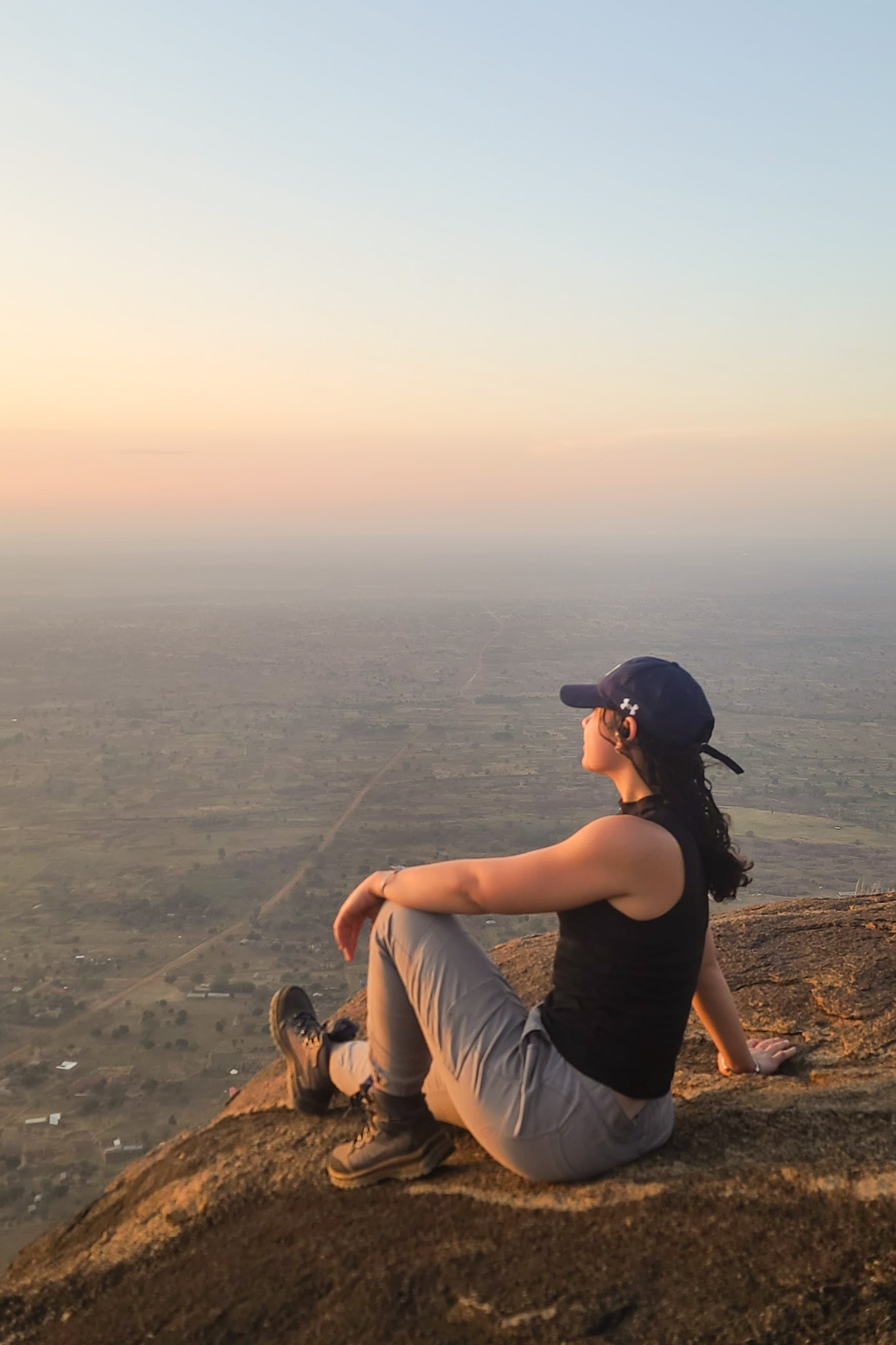 A person in a cap sits on a rock, overlooking a vast landscape at sunrise, with a serene sky blending into warm earthy tones.