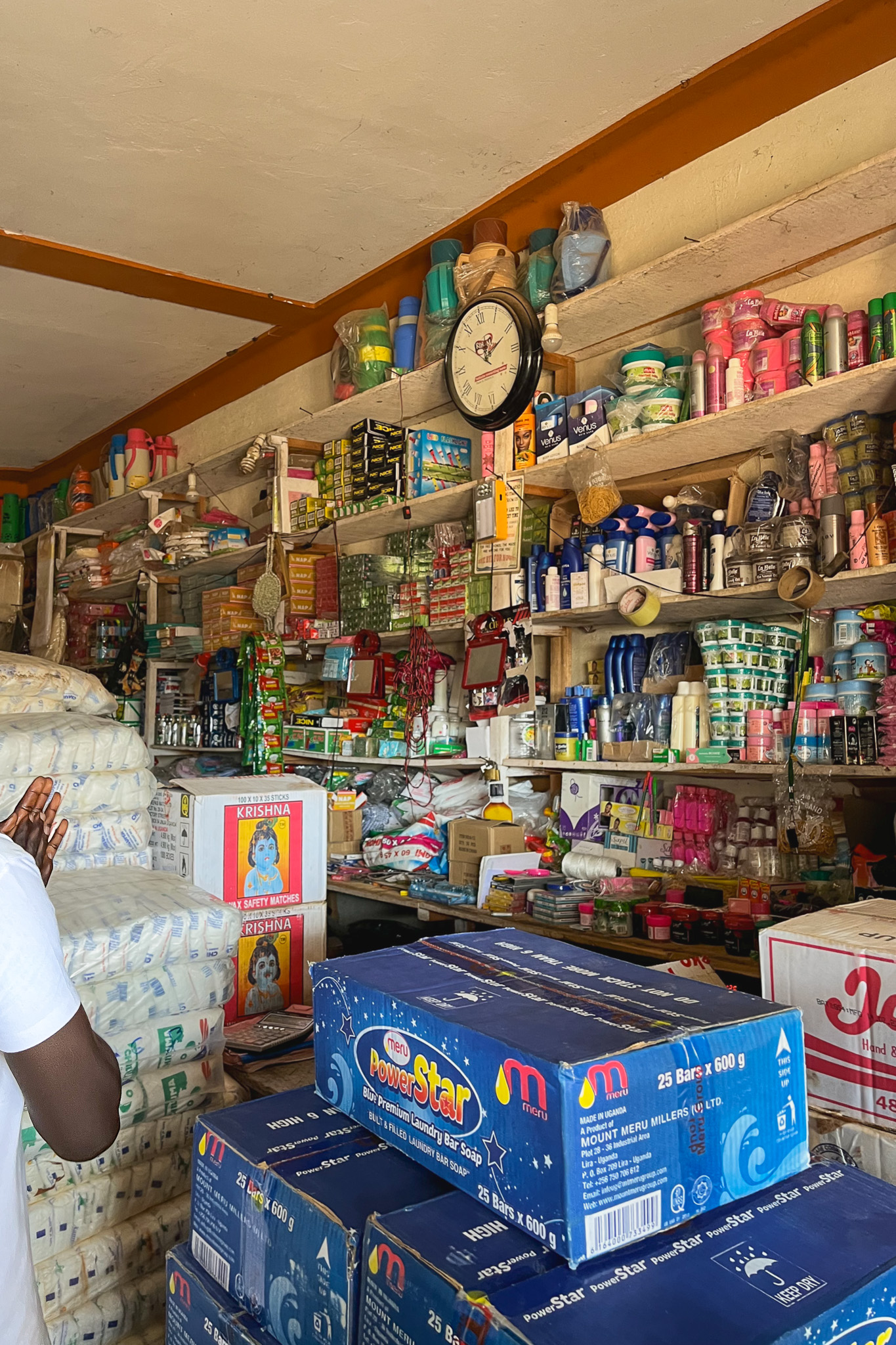 Crowded shop interior with shelves filled with household goods, toiletries, and piles of laundry soap boxes. A wall clock and packages are visible.