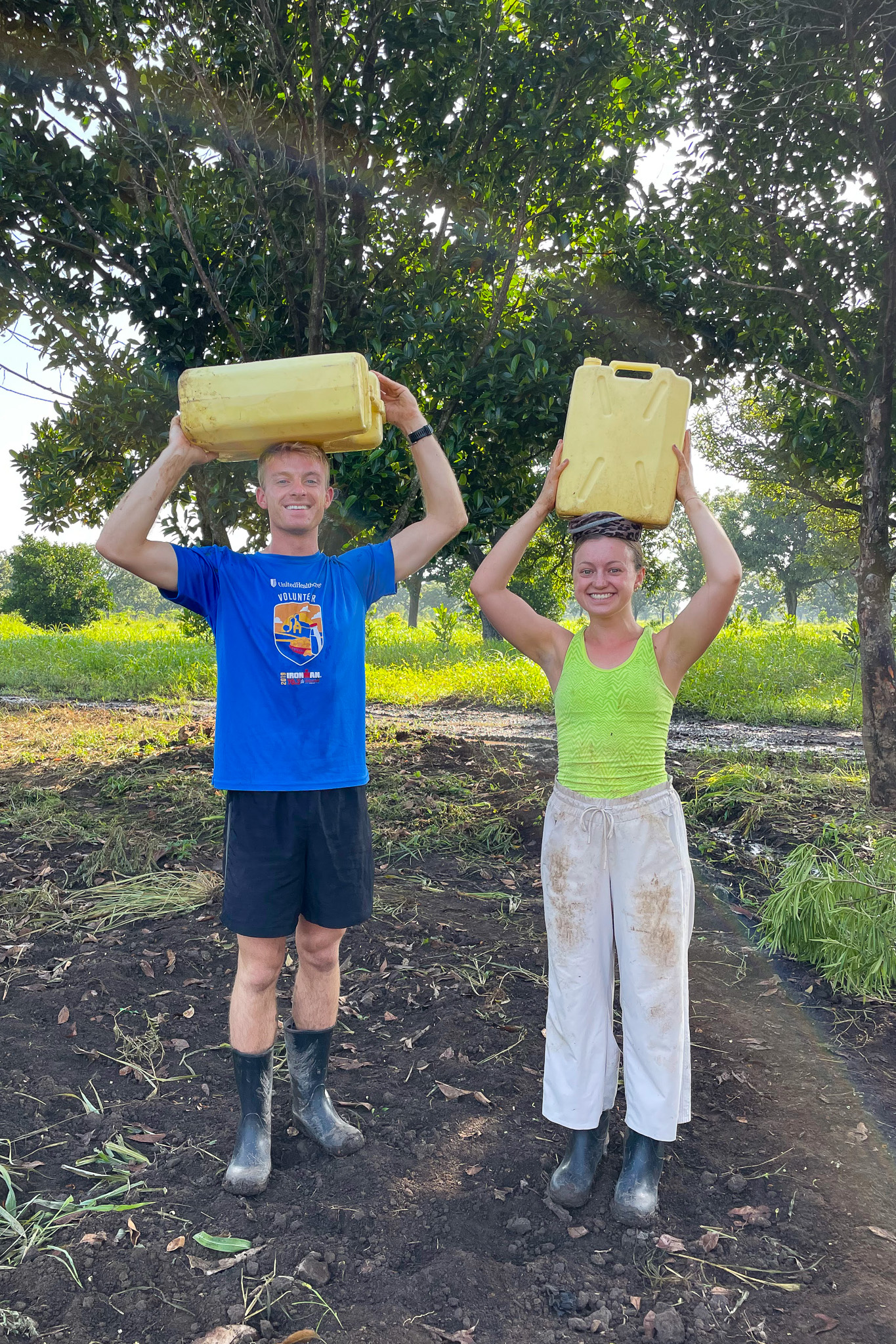 Two people smiling, carrying yellow jerry cans on their heads, standing on soil with trees in the background.