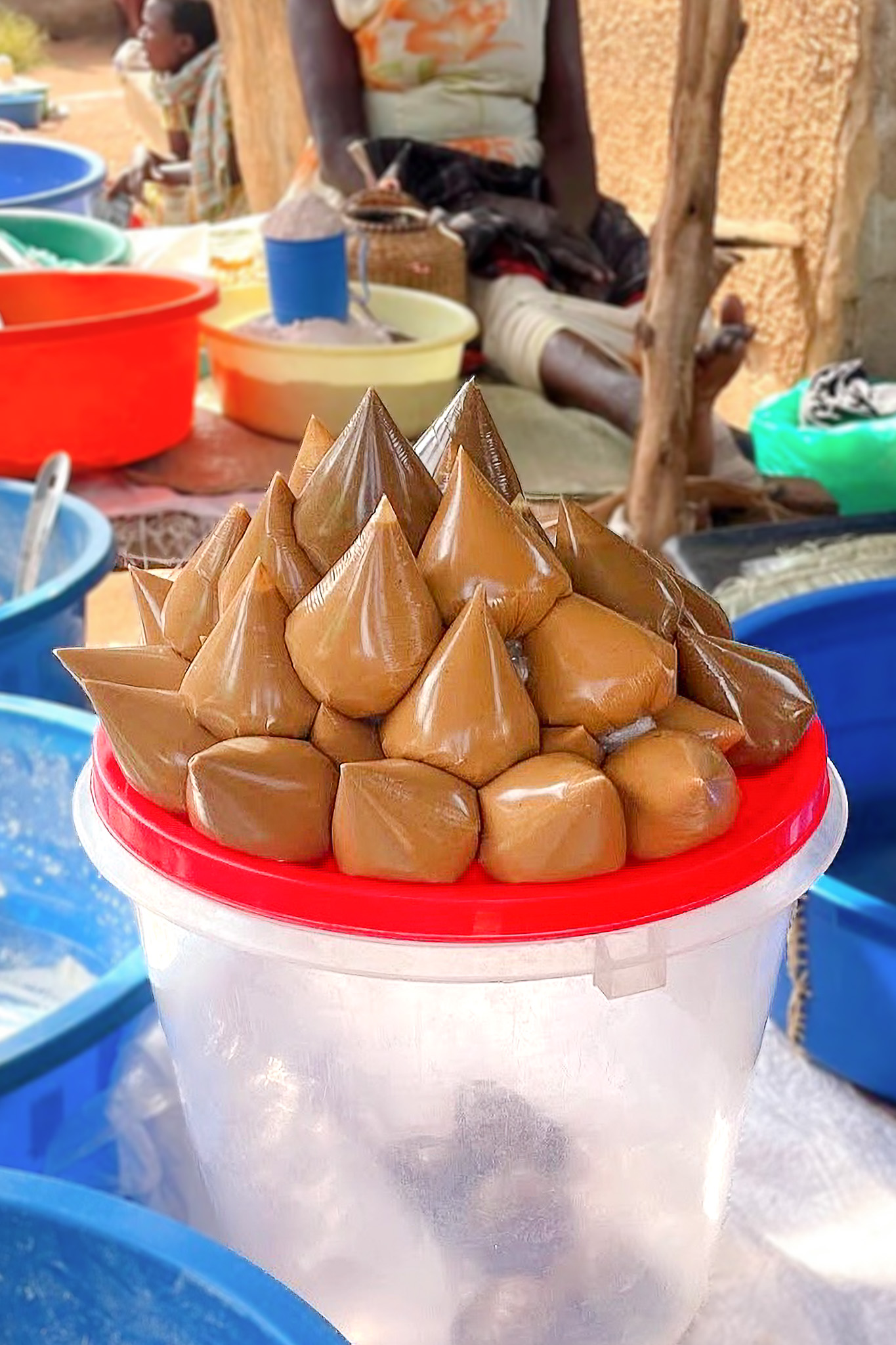 Small conical bags of peanut paste arranged on a red lid atop a plastic container at a market stall.