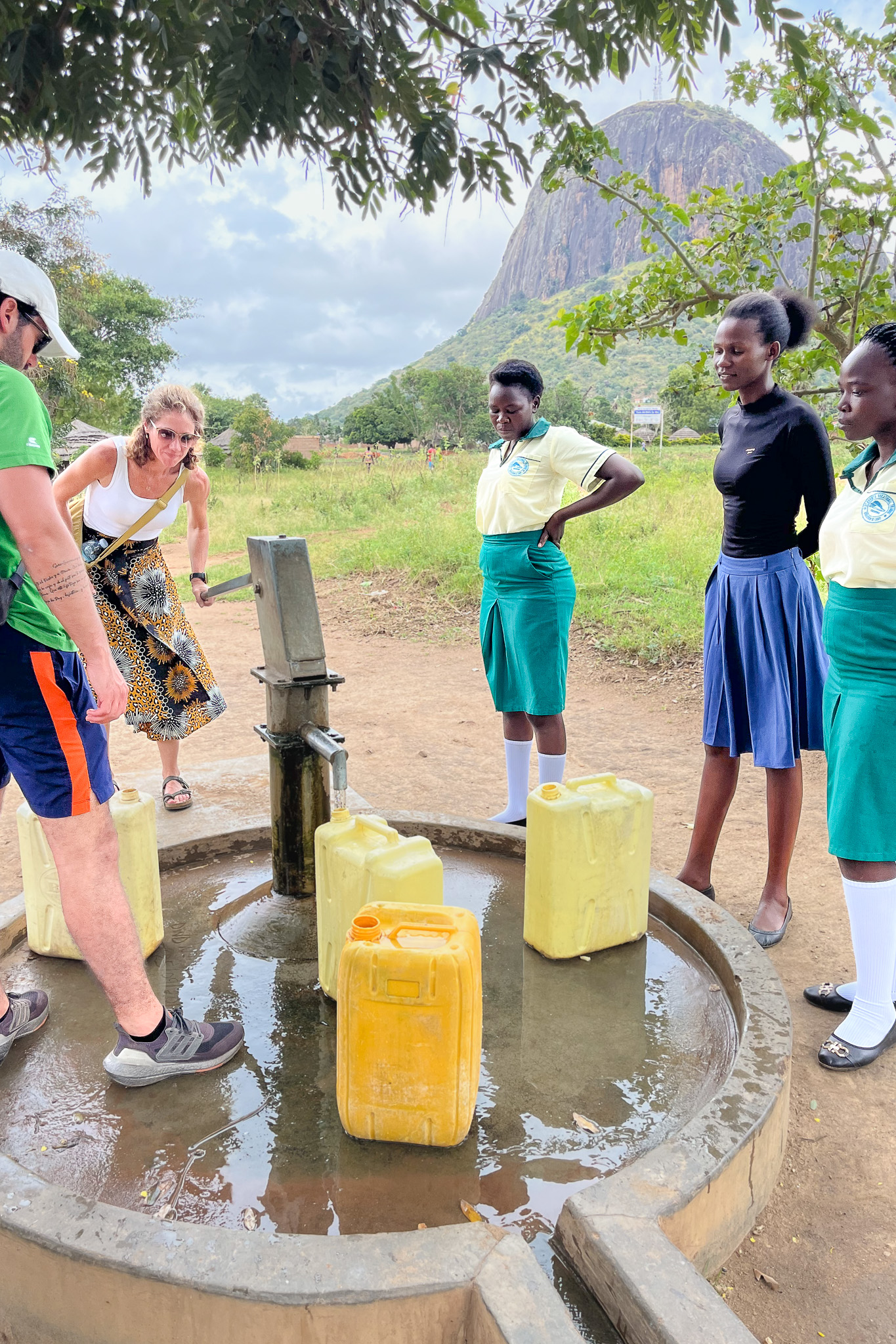 People gather around a water pump with yellow jerry cans, set against a backdrop of a mountain and greenery.