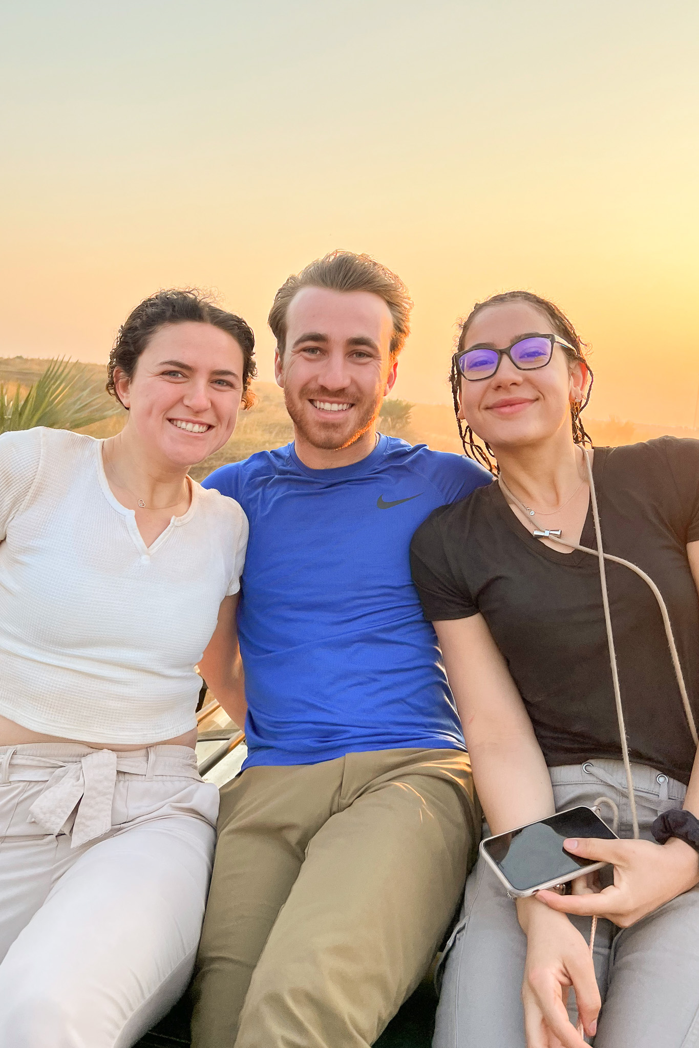 Three people smiling and sitting together outdoors at sunset, with a soft orange sky in the background.