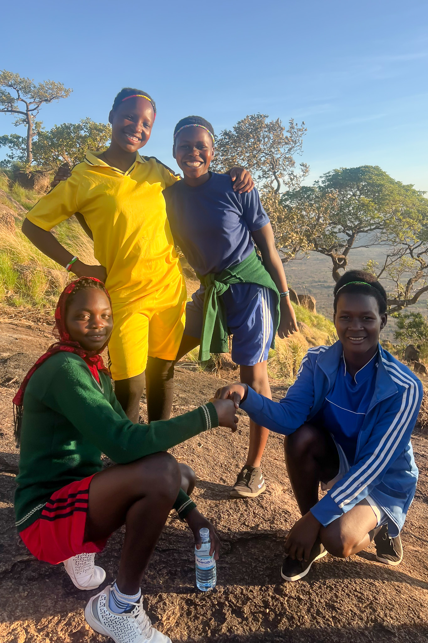 Four friends smiling and posing on a rocky hillside, wearing colorful athletic clothing, with trees and a clear blue sky in the background.