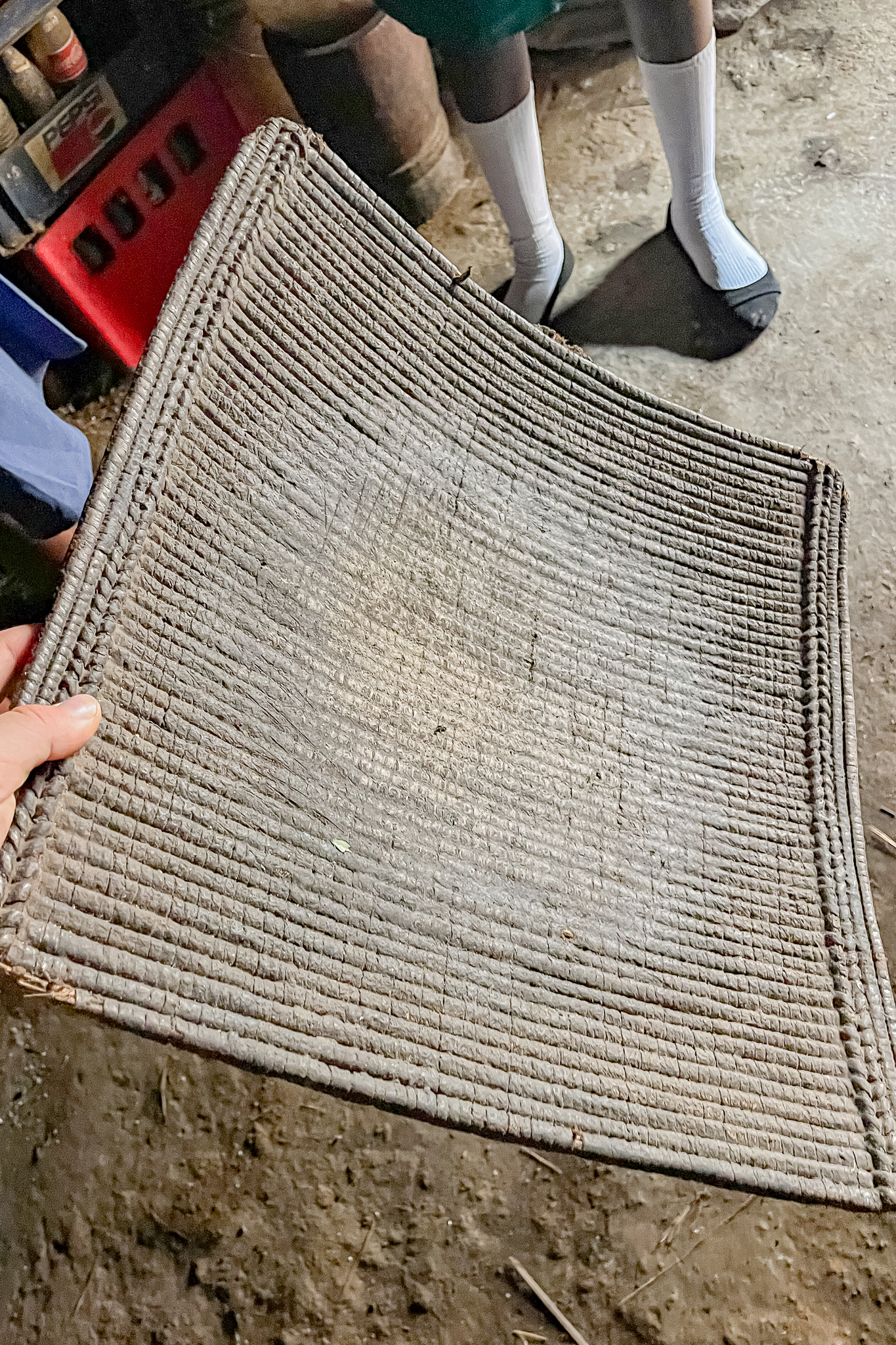 Person holding a woven mat indoors, with two visible legs wearing white socks and sandals in the background.