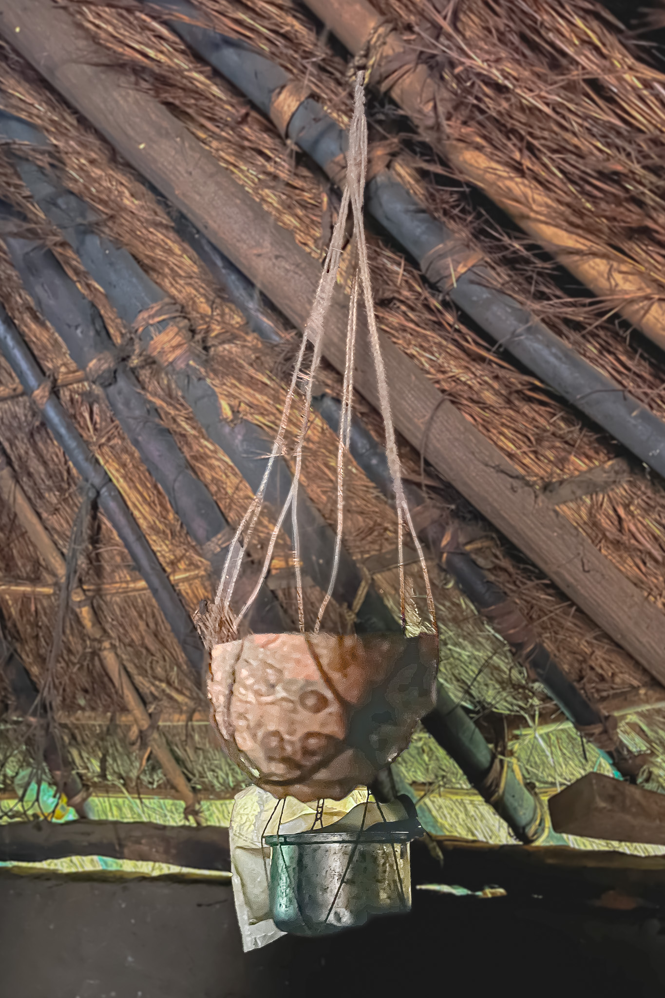 A handmade pottery object suspended from a thatched roof by strings, with a rough textured surface and small metallic cups hanging below.