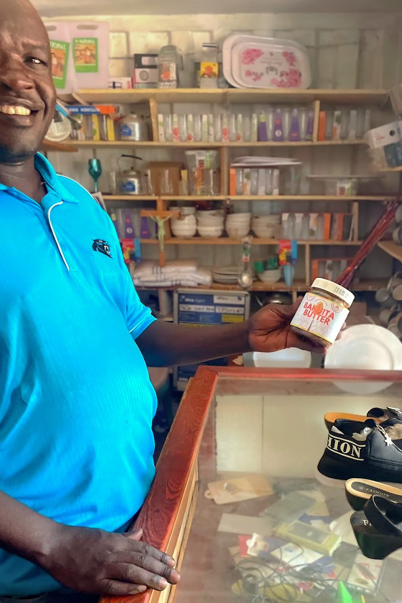 Smiling man in a blue shirt holds a jar of "Bakita Butter" in a store filled with assorted household items on shelves.