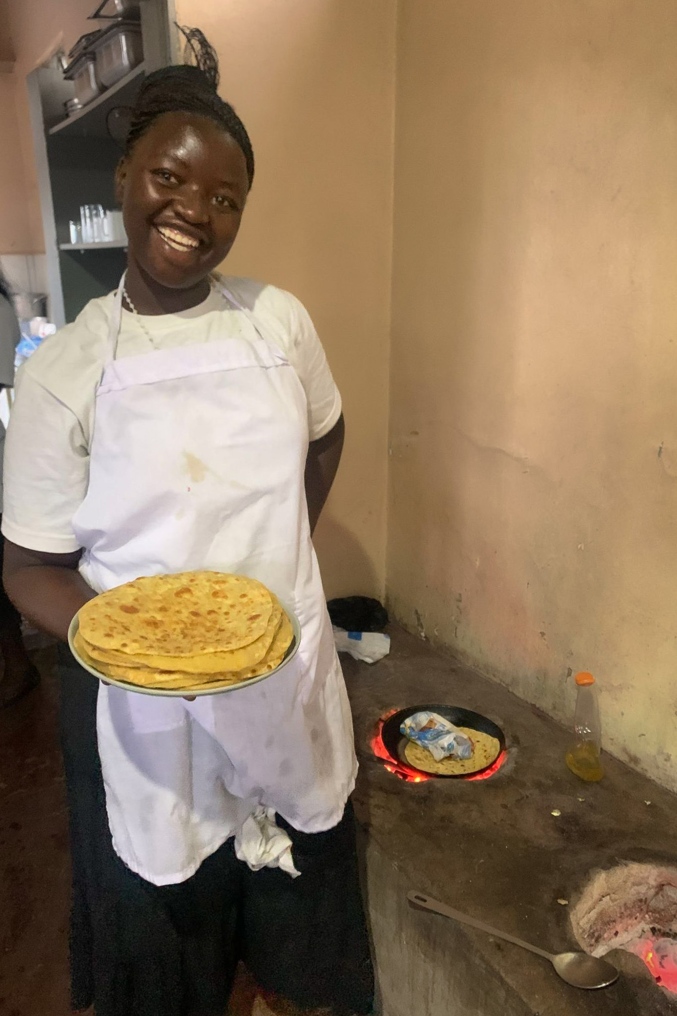 Smiling woman in a white apron holding a plate of freshly made chapatis in a rustic kitchen.