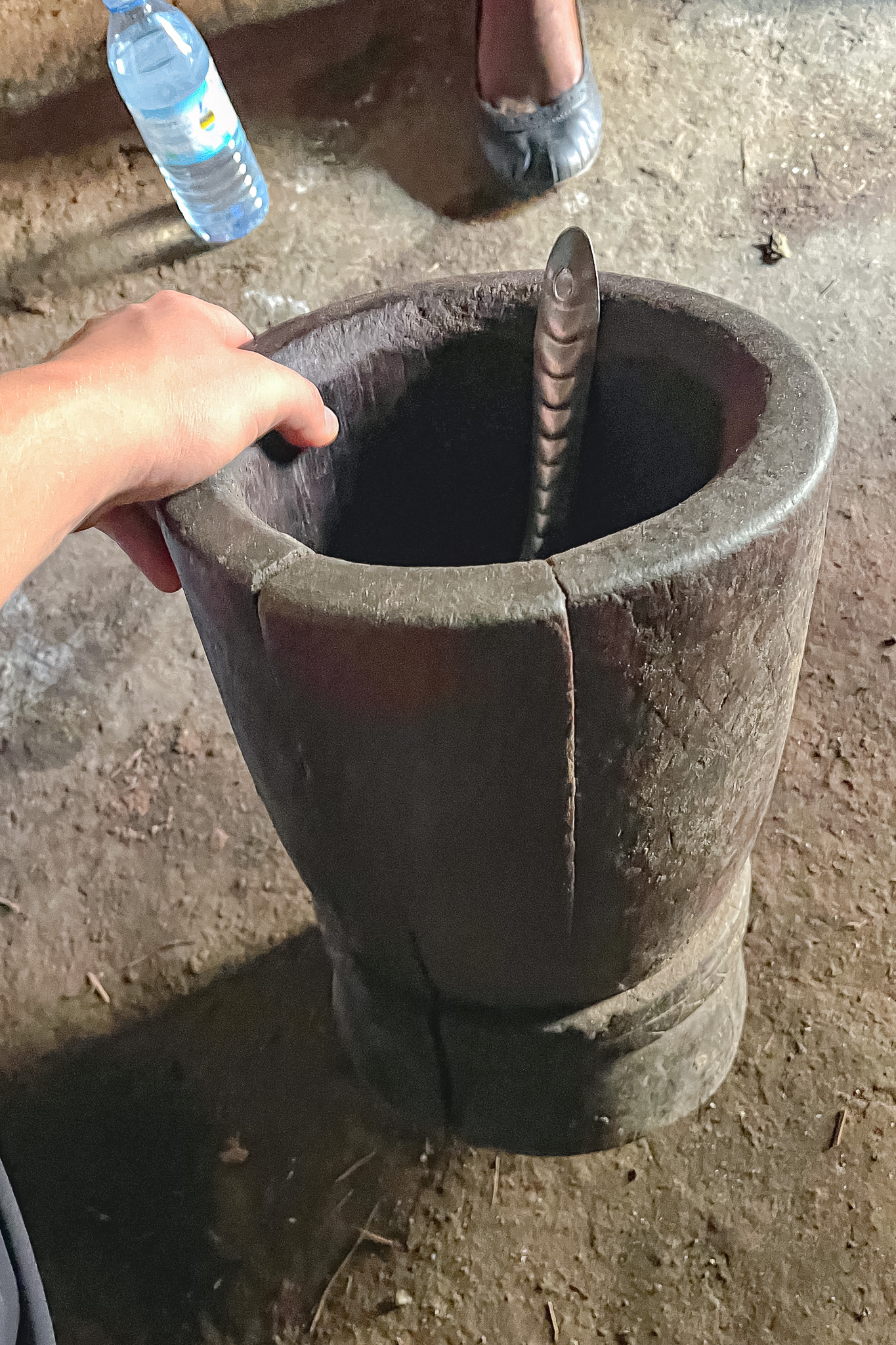 A hand touches a large wooden mortar with a metal pestle inside, on a dirt floor. Nearby is a water bottle and a person's foot in a shoe.