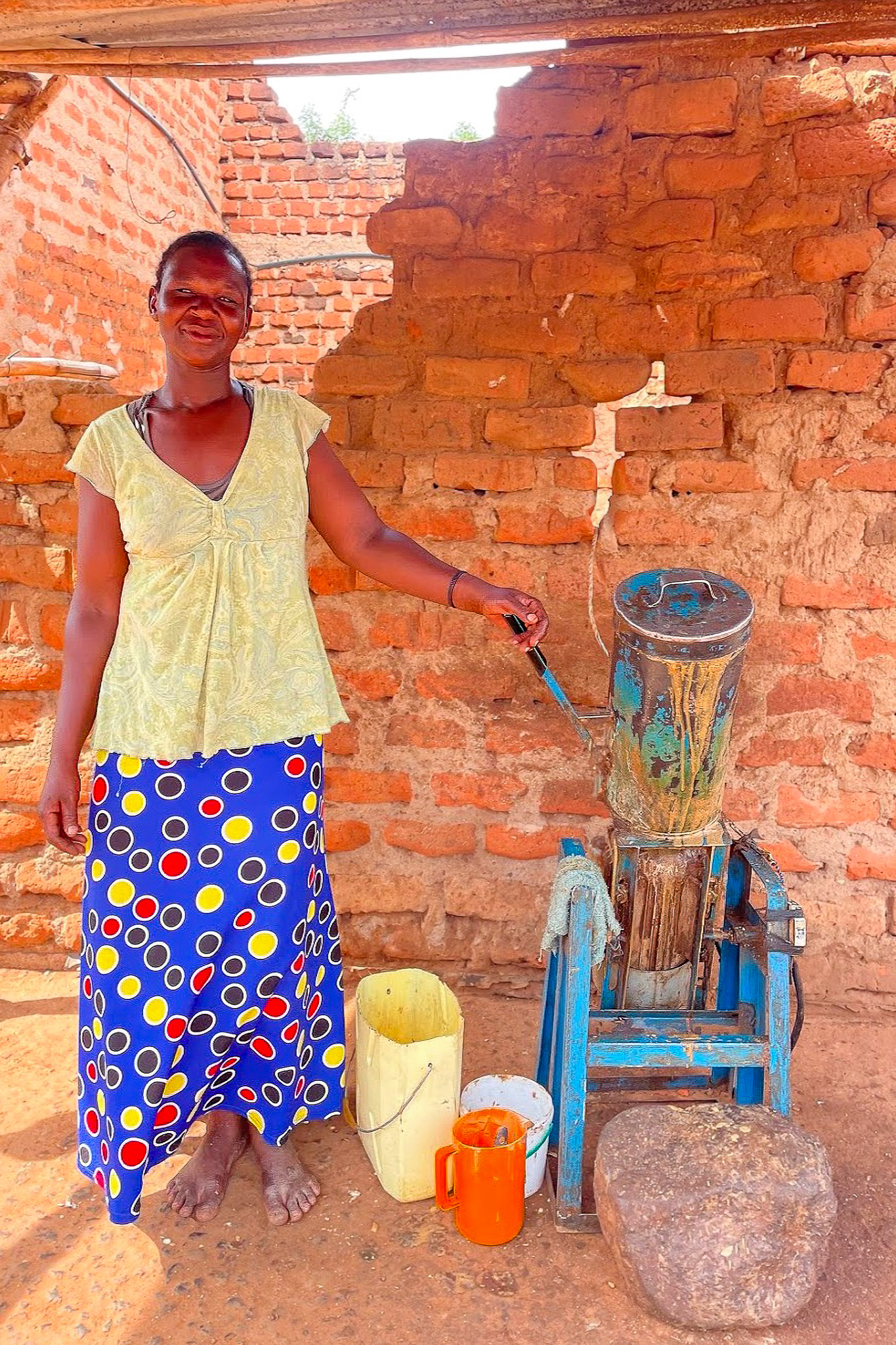 Woman smiling beside a manual sugarcane juicer in front of a brick wall, with colorful containers below.
