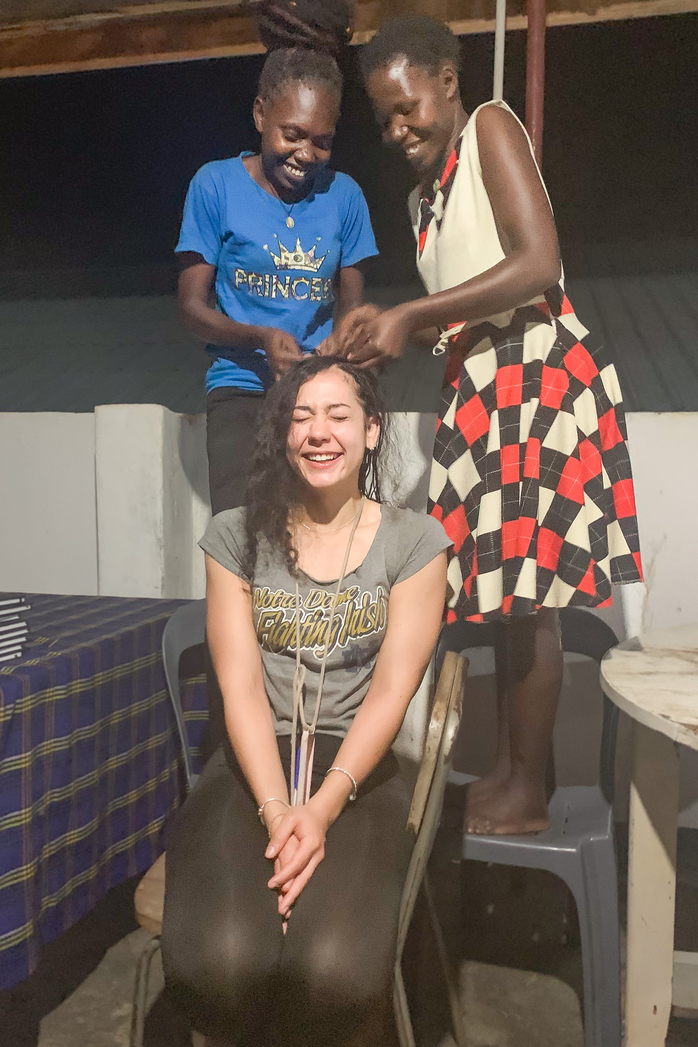 Two women braid another woman's hair as she sits, smiling with closed eyes, on a chair indoors.