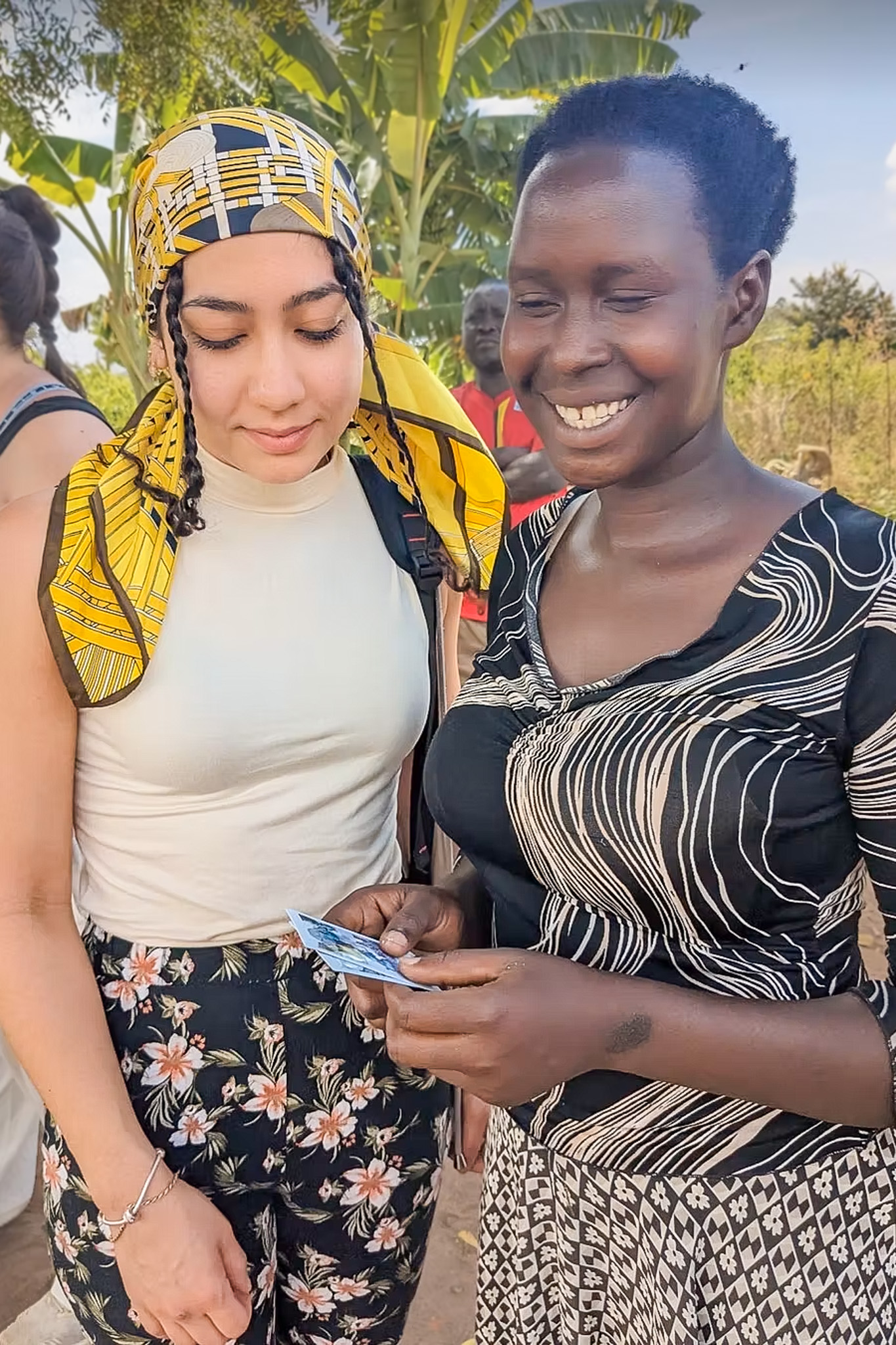 Two women standing together outdoors, one wearing a yellow patterned headscarf, the other holding a small photo, both smiling, with greenery in the background.