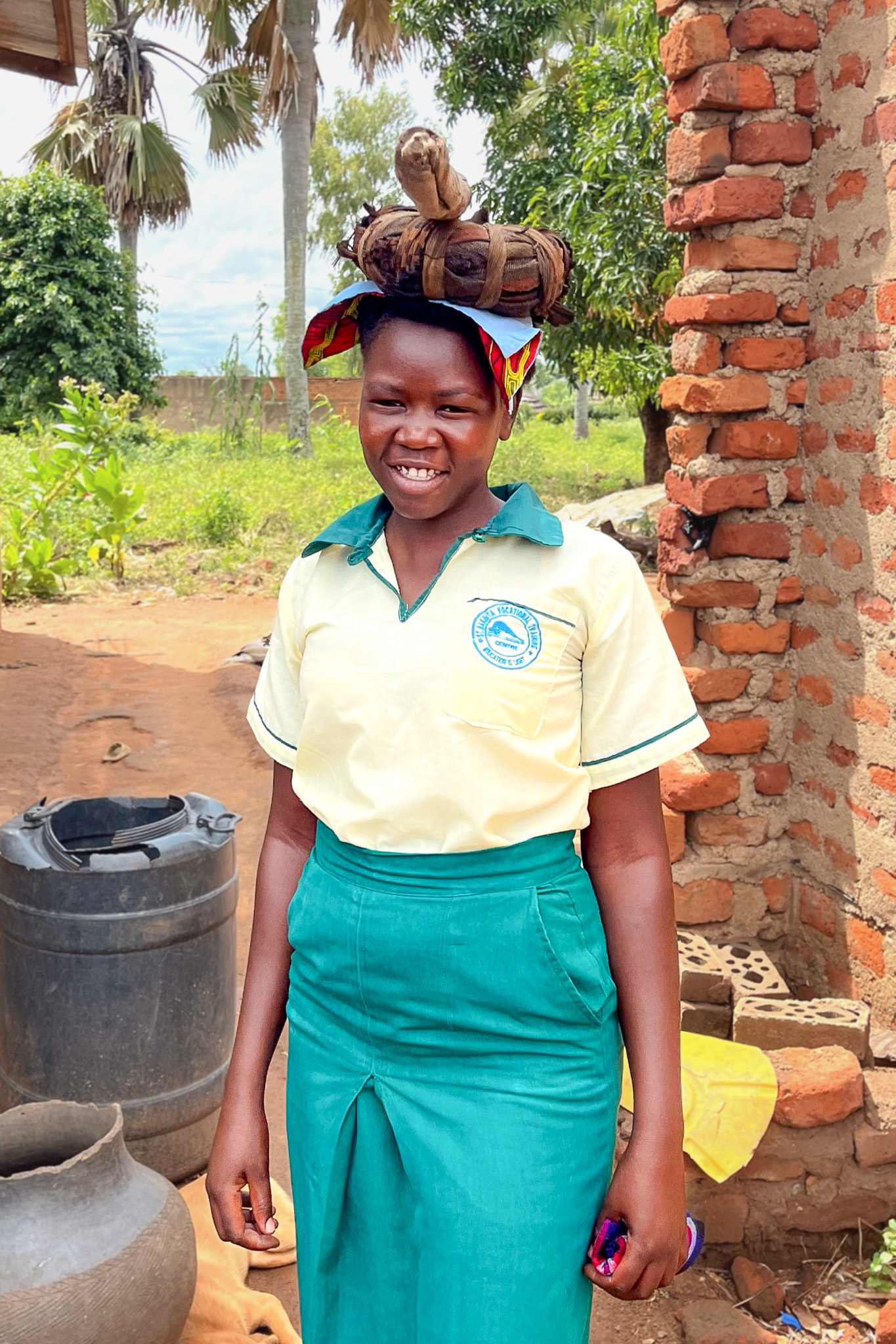 Smiling woman in a school uniform carries a bundle on her head, standing outside near brick building and greenery.