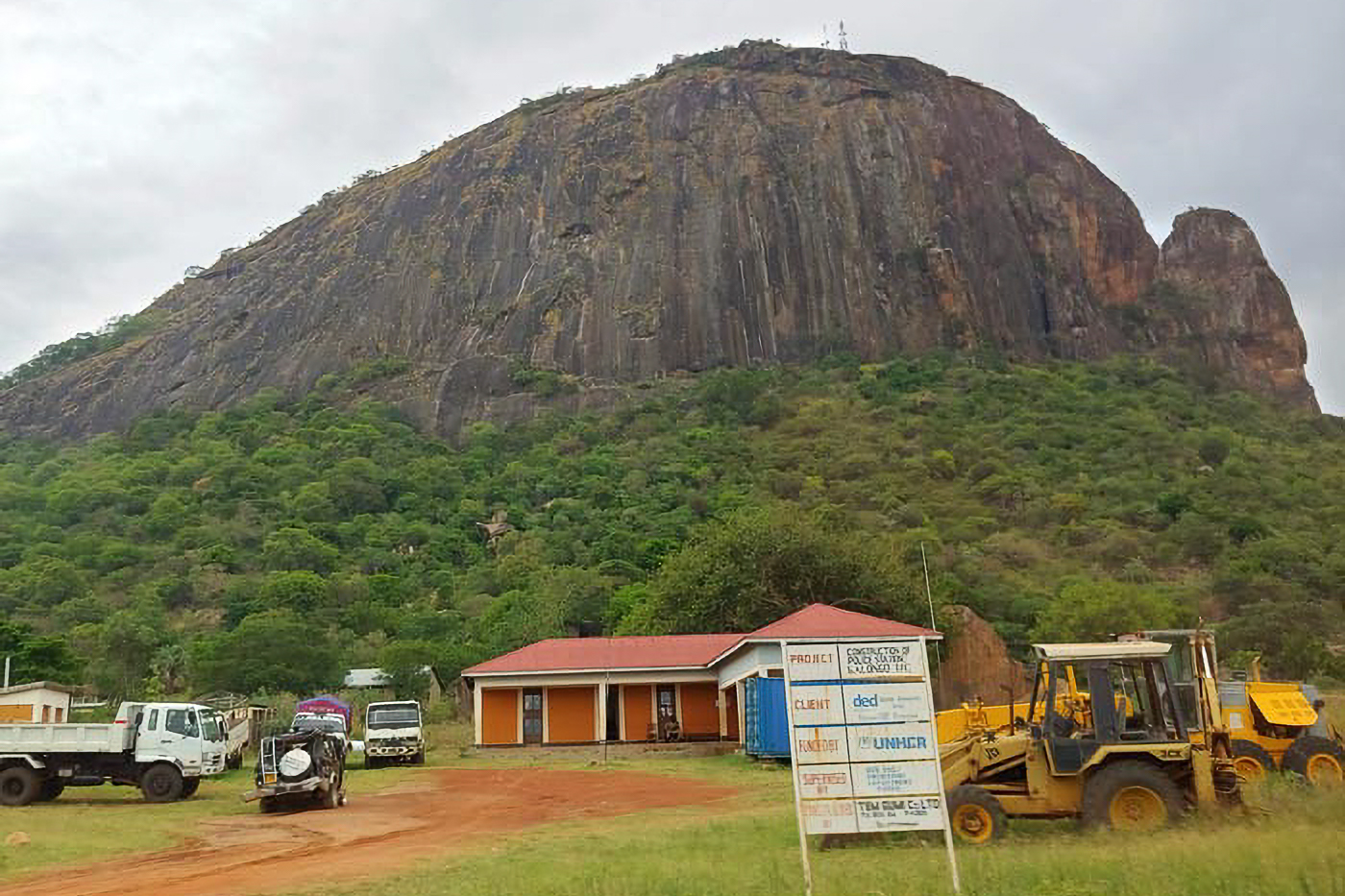 A large rocky hill with a small building, vehicles, and construction equipment in the foreground under a cloudy sky.