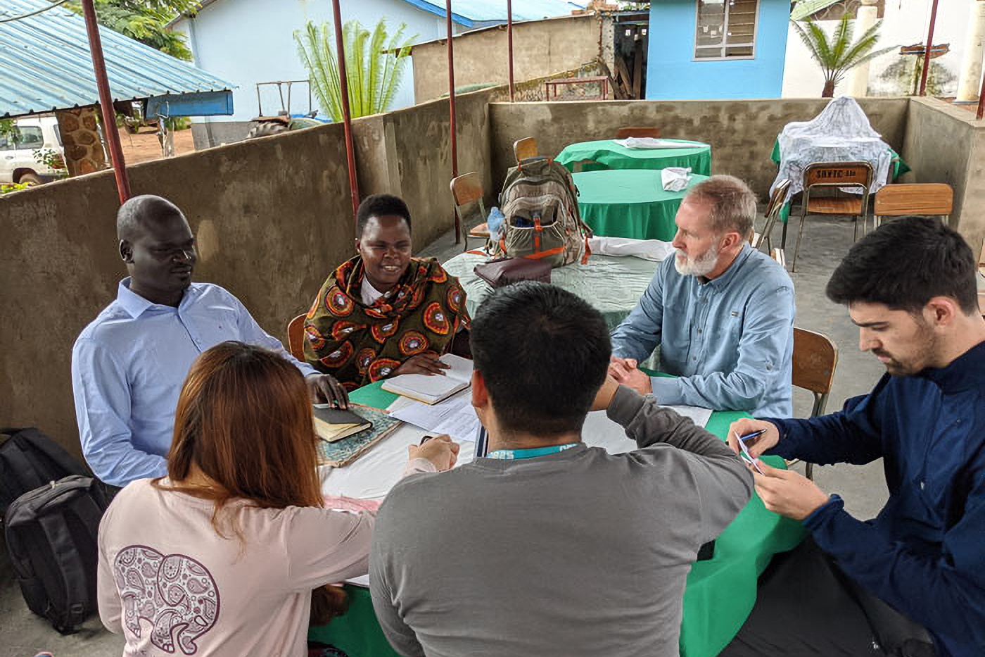 A diverse group of people sitting around a table in discussion on an outdoor patio with green tablecloths and backpacks nearby.