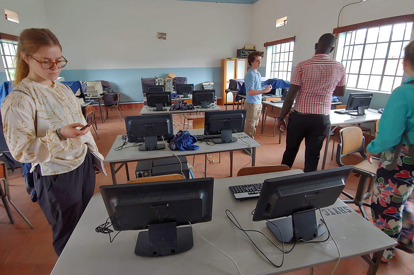 People exploring a computer lab in a classroom with several desks and monitors. One person is using a phone, and others are arranging equipment.