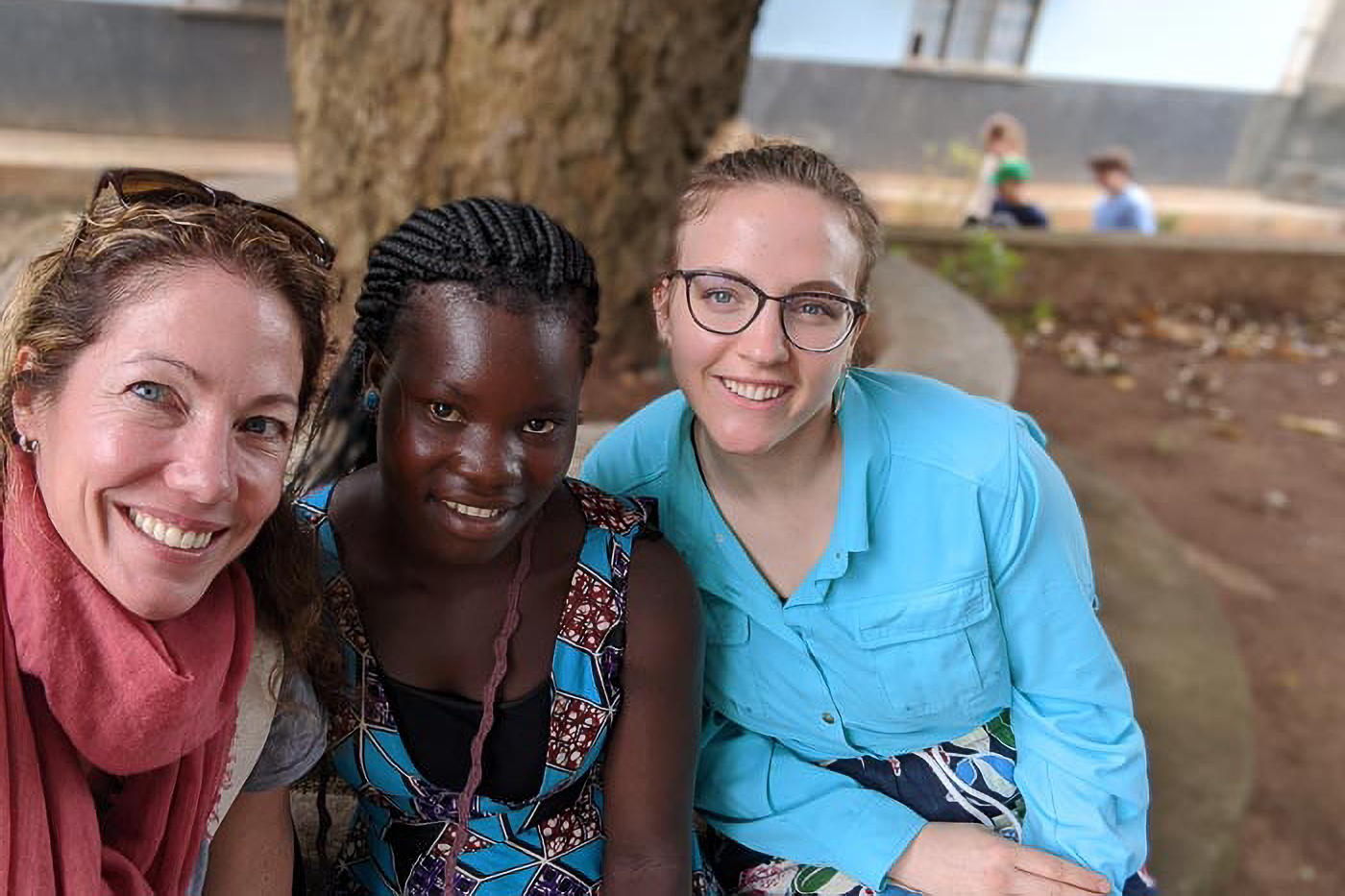 Three women smiling and sitting together outdoors, with a large tree trunk in the background.