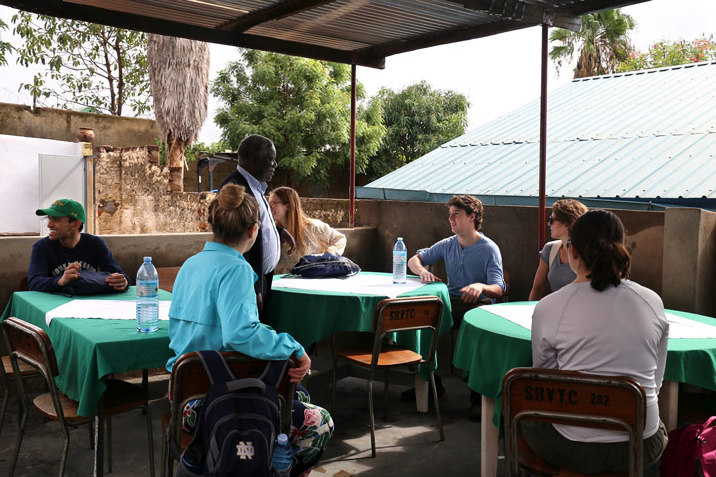 People sitting and standing around tables with green tablecloths in an outdoor setting, engaged in conversation, with trees and a corrugated roof visible.
