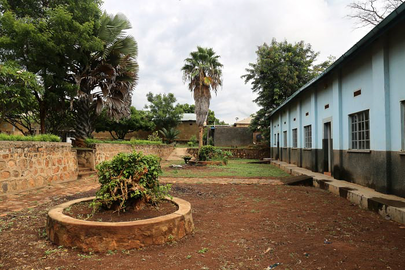 Outdoor courtyard with a circular stone planter, trees, and a blue building with multiple windows on the right. Cloudy sky above.