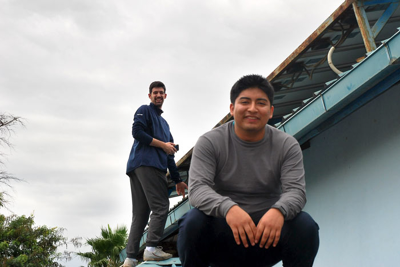 Two men on a rooftop, one crouching and smiling, the other standing and smiling, with a cloudy sky in the background.
