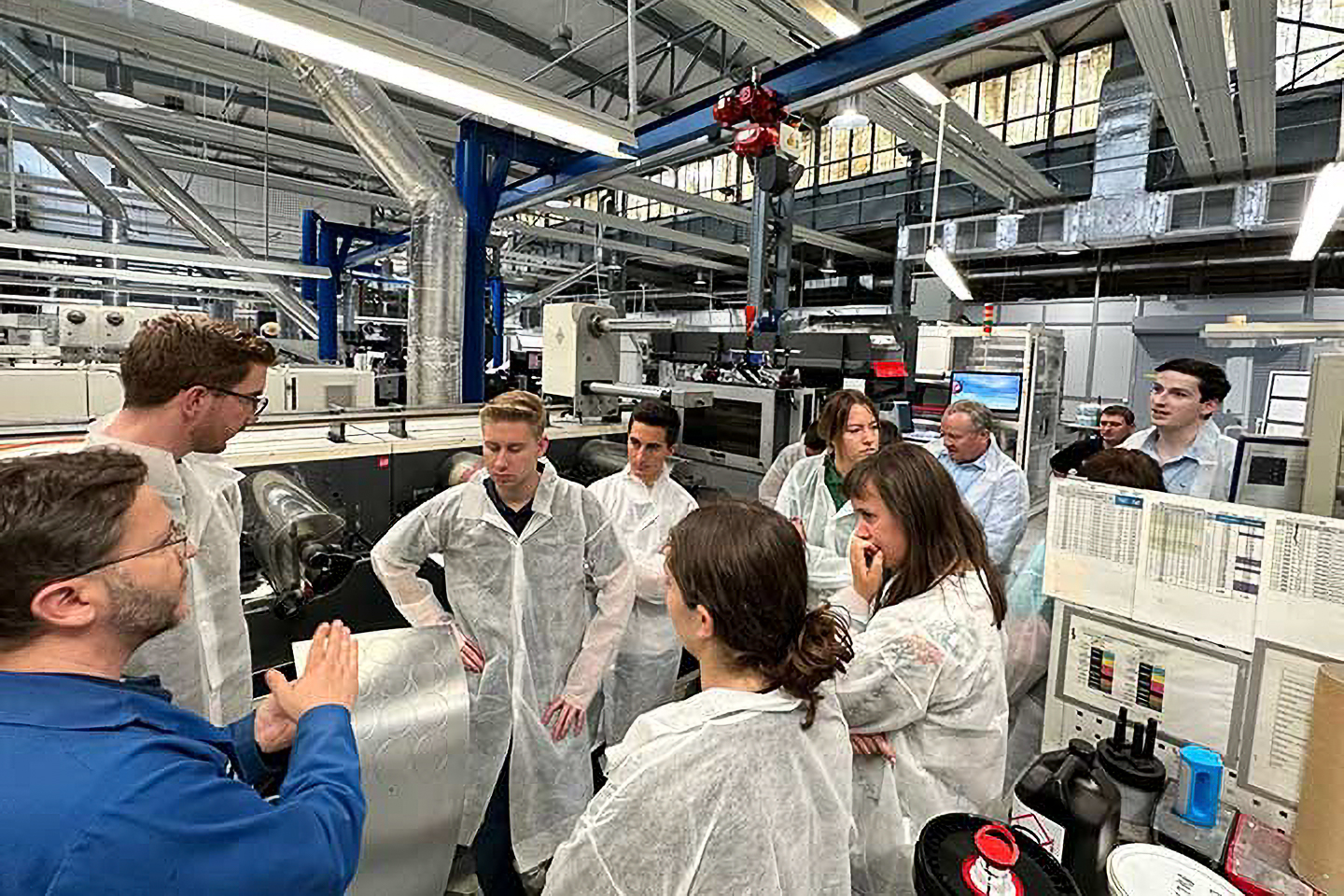 A group of people in protective clothing listen to a guide in an industrial workshop filled with machinery and equipment.