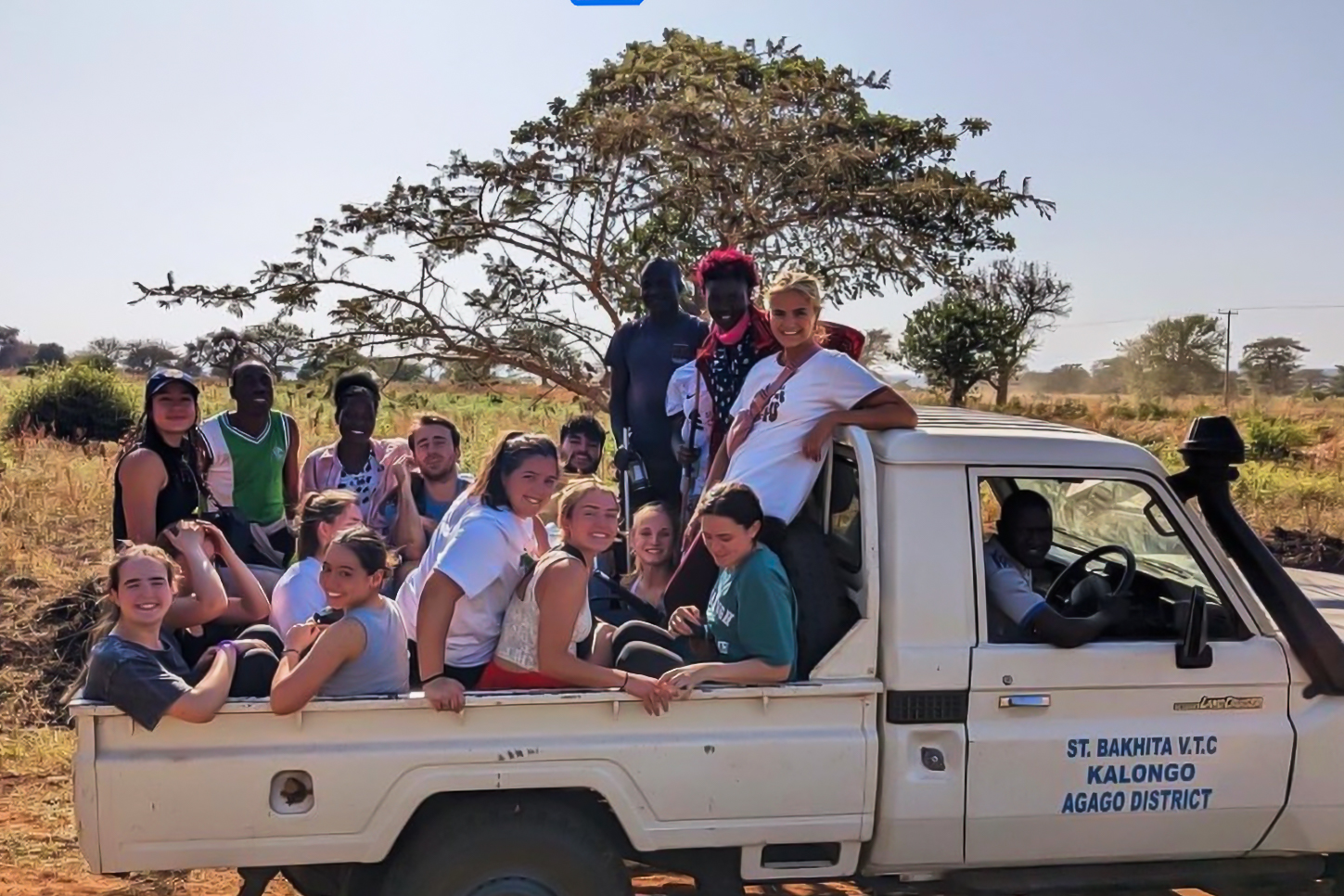 A group of people sitting in and on a truck labeled "St. Bakhita V.T.C, Kalongo, Agago District," against a rural backdrop with trees.